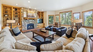 Un salon chaleureux avec une cheminée en pierre, des étagères en bois, des sièges confortables et de grandes fenêtres donnant sur l’extérieur.