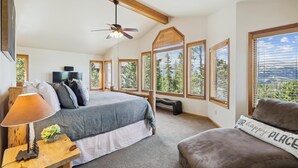 Une chambre à coucher avec un lit, un bureau, une lampe et une vue sur les arbres, visible à travers de grandes fenêtres.