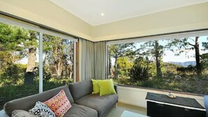 Ein modernes Wohnzimmer mit einem grauen Sofa, einem kleinen Tisch und einem großen Fenster mit Blick auf Bäume und Hügel.