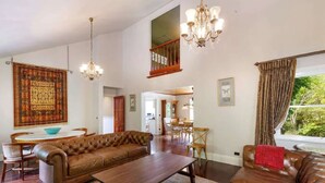 A spacious living room with a high ceiling, a chandelier, a brown leather sofa, a dining area, and a view of greenery outside.