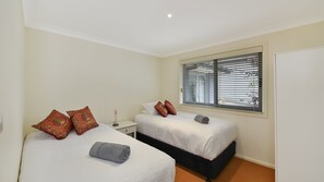 A bedroom with two beds, each with a white bedspread and decorative pillows, a nightstand, a bedside table, and a window with blinds. - Aisling View in Blackheath (Blackheath)