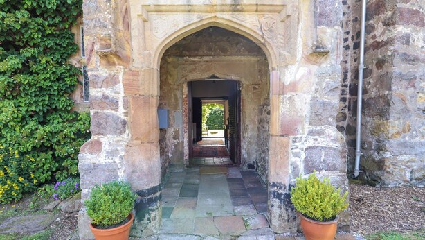 A stone archway leading to a courtyard with a gravel path, surrounded by greenery and potted plants. - Y Fferm (Mold)