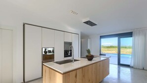 Una cocina moderna con una isla central, electrodomésticos integrados y un amplio ventanal corredero que da a una piscina.