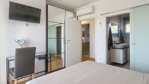 Un dormitorio moderno con cama, escritorio, silla, televisor, espejo y vestidor.