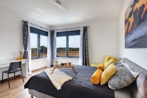 Une chambre avec un grand lit, un bureau et une chaise. Il y a deux fenêtres avec des rideaux, un tableau au mur et une vue sur la campagne.