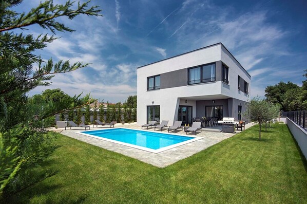 Une maison moderne à deux étages, dotée d’une piscine, d’un espace salon extérieur et d’une pelouse bien entretenue.