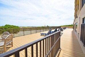 Um deck de madeira com espreguiçadeiras e uma pequena mesa, com vista para uma praia e um cercado.
