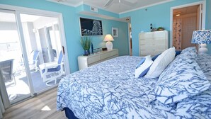 Quarto com uma cama grande, um ventilador de teto, uma escrivaninha com luminária e uma porta de vidro deslizante que dá para uma varanda com cadeiras.