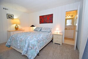 A bedroom with a bed, two bedside tables, a nightstand, a wall-mounted artwork, and a door leading to a bathroom. - Alameda 5 2 Bedroom Condo by RedAwning (Ocean City)