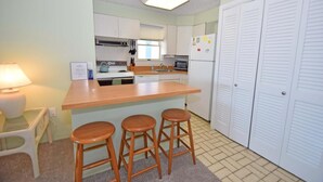 A compact kitchen with a central island, wooden stools, a microwave, a refrigerator, and a window with blinds. - Alameda 5 2 Bedroom Condo by RedAwning (Ocean City)