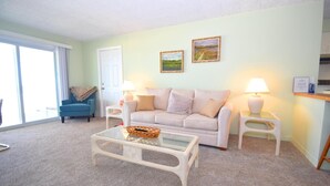 A living room with a light-colored sofa, two side tables with lamps, a glass coffee table, and a blue armchair. - Alameda 5 2 Bedroom Condo by RedAwning (Ocean City)
