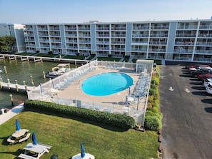 A swimming pool with lounge chairs and umbrellas, surrounded by a fence, with a parking lot and a building in the background. - Thunder Island 16a 2 Bedroom Condo by RedAwning (Ocean City)