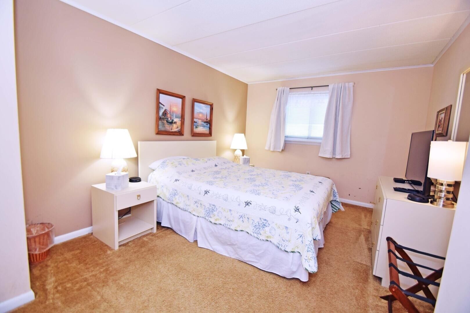 Une chambre à coucher comprenant un lit, deux tables de chevet avec des lampes, un bureau, une chaise, une fenêtre avec des rideaux et deux tableaux encadrés accrochés au mur.