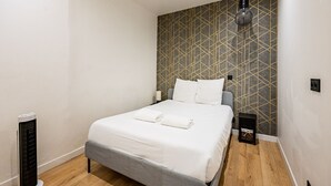 Ein ordentlich bezogenes Bett in einem Zimmer mit Wand im geometrischen Muster, ein Heizkörper und ein kleiner, schwarzer Lautsprecher auf dem Boden.