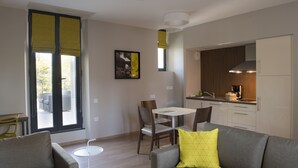 A modern living room with a grey sofa, a yellow pillow, and a small white table. There is a dining area with a white table and chairs, and a kitchen with wooden cabinets and a stainless steel range hood. - Le Château des Plans (Parthenay)