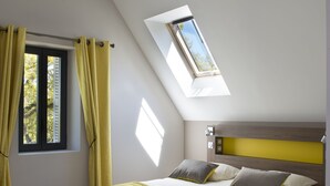 A bedroom with a skylight, a bed with pillows, a chair, and a window with curtains. - Le Château des Plans (Parthenay)
