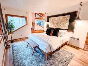 A bedroom with a large bed, a wooden bench, a potted plant, a map on the wall, and a window with a view of trees. - Private Cabin, Pets Welcome, Hot Tub surrounded by nature (Gatlinburg)