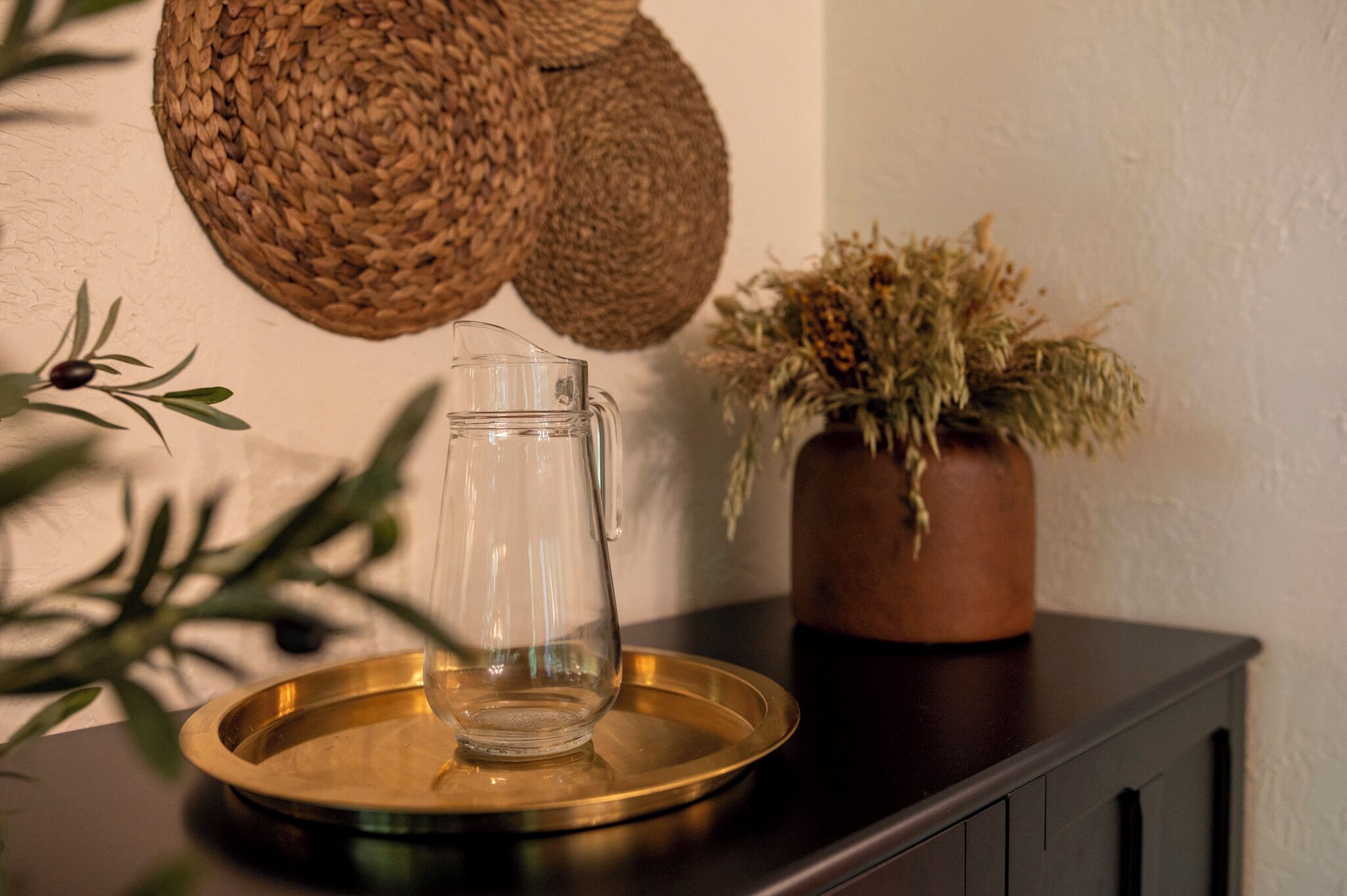 Ein klarer Glasbehälter auf einem goldenen Tablett, ein gewebter Korb und ein Topfpflanzenarrangement mit getrockneten Blumen auf einer dunklen Holzoberfläche.
