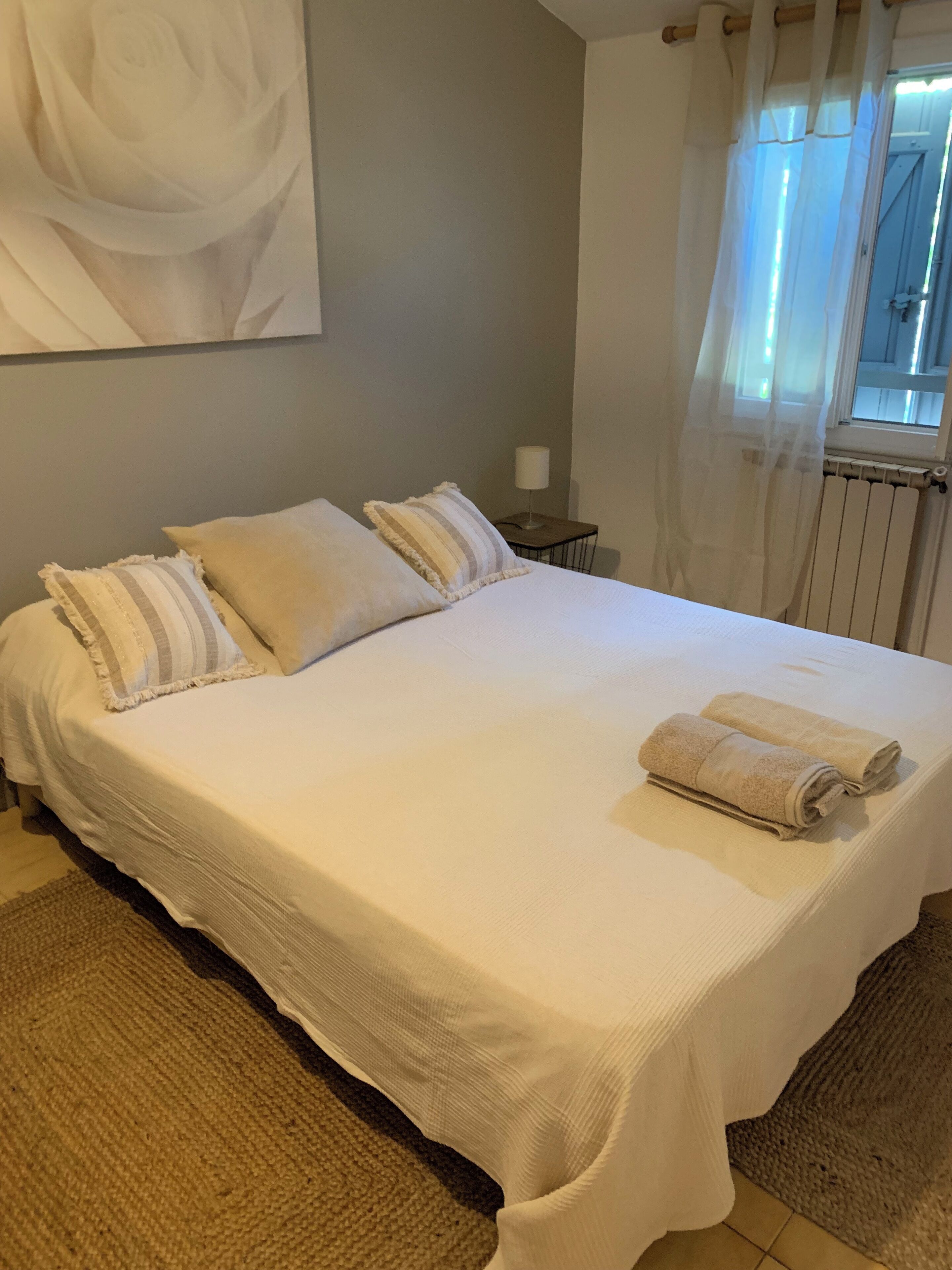A neatly made bed with white linens and striped pillows, a bedside lamp, and a window with sheer curtains.