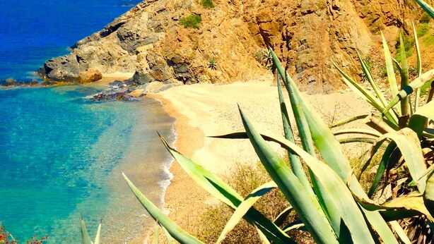 Paesaggio costiero con una collina rocciosa, mare blu cristallino e spiaggia sabbiosa.