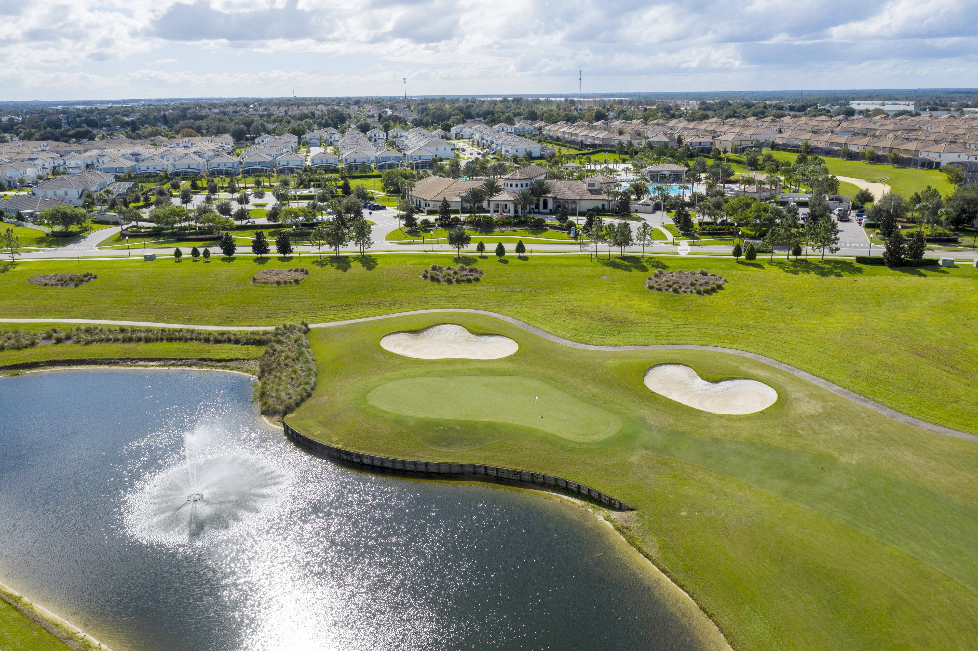 Vue aérienne d’un parcours de golf avec des bunkers de sable, un étang et un quartier résidentiel en arrière-plan.