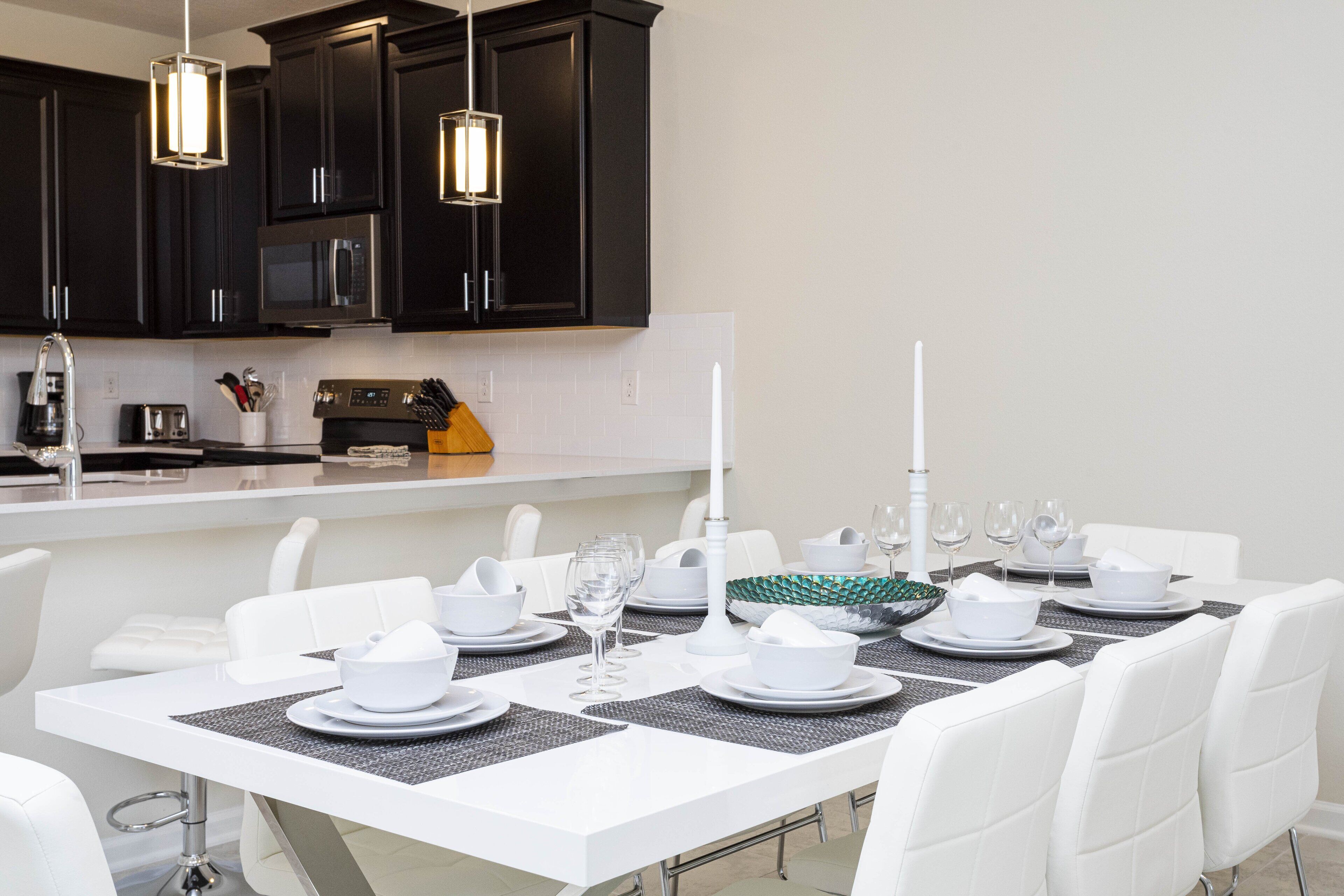A modern kitchen with dark cabinets, a microwave, and a dining area set with white chairs, a white table, and a black placemat.