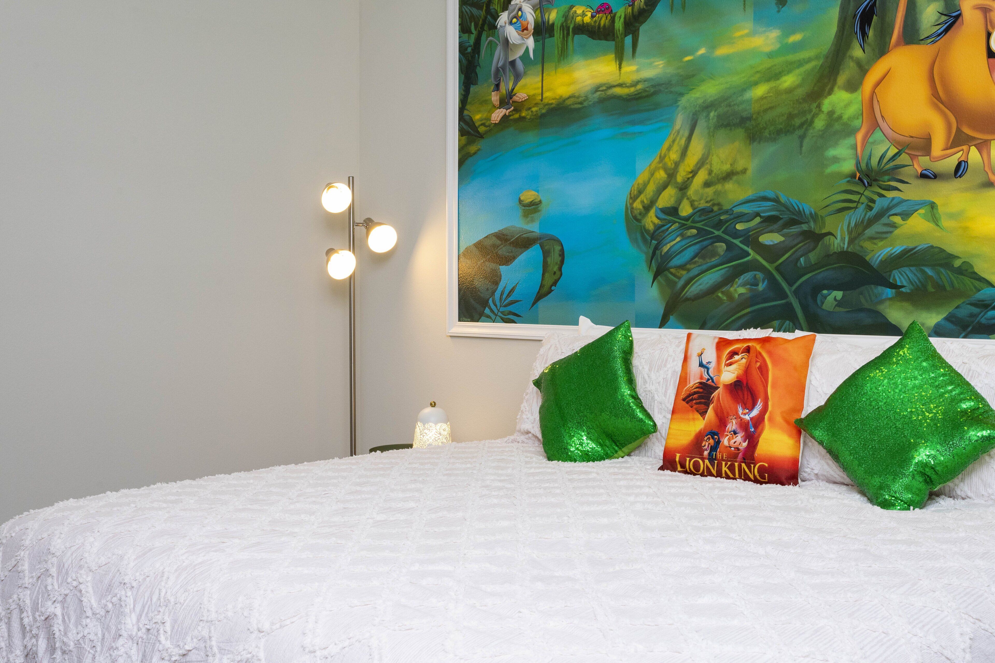 Una cama con cubre sábana blanco texturizado, dos almohadas verdes, un cartel de 'El Rey León' en la pared y una lámpara de pie con tres luces.