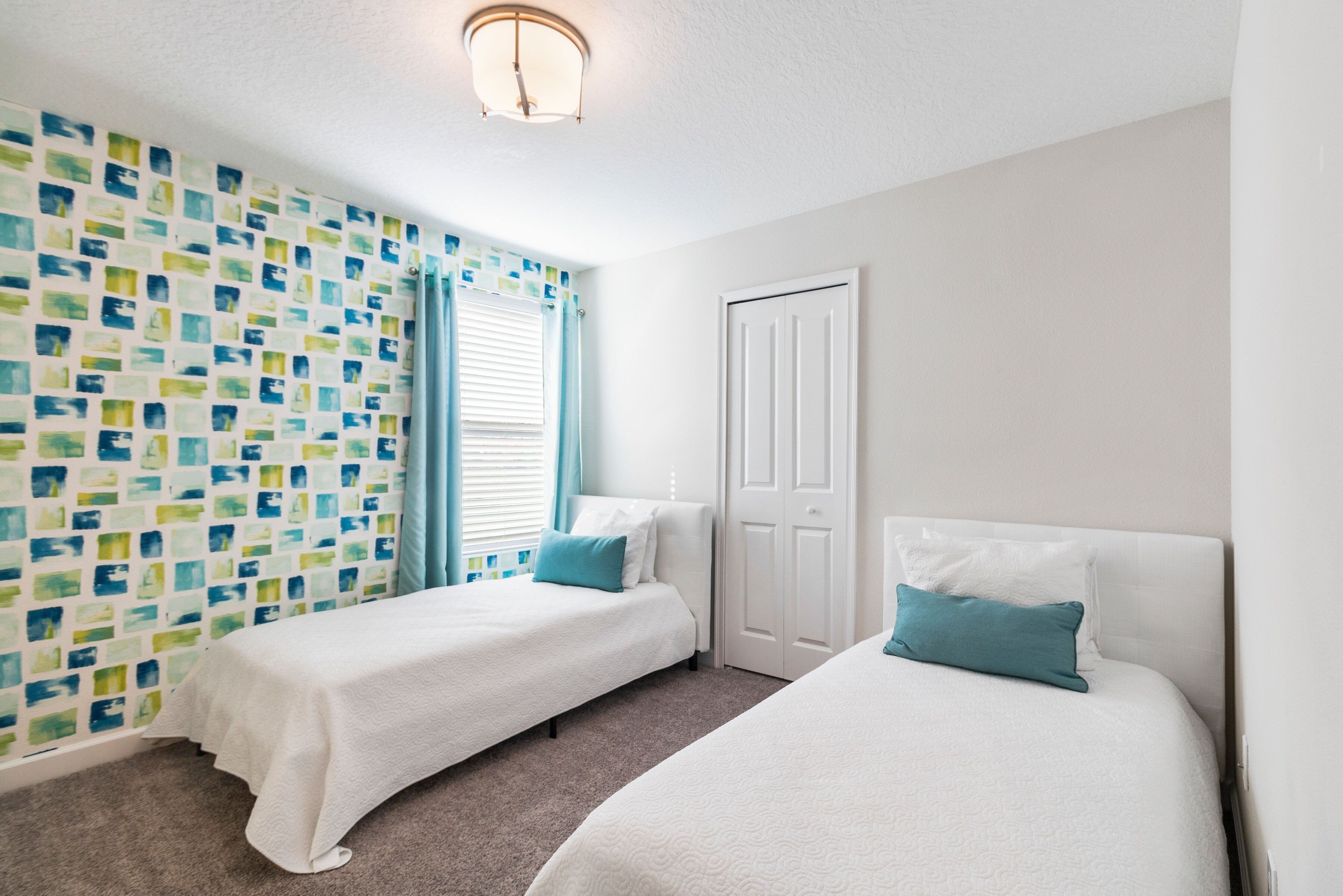 A room with two single beds, a white door, a patterned wall, and a window with blue curtains.