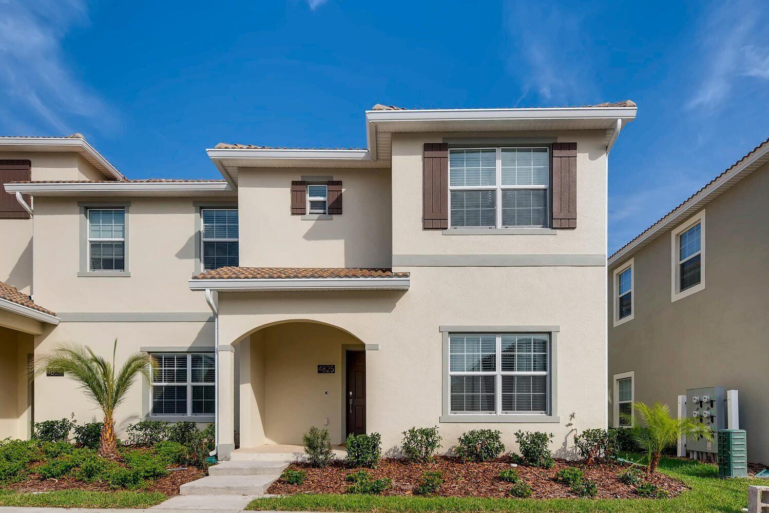 A two-story residential building with a beige facade, a central entrance with an archway, and a small front garden with palm trees and shrubs.