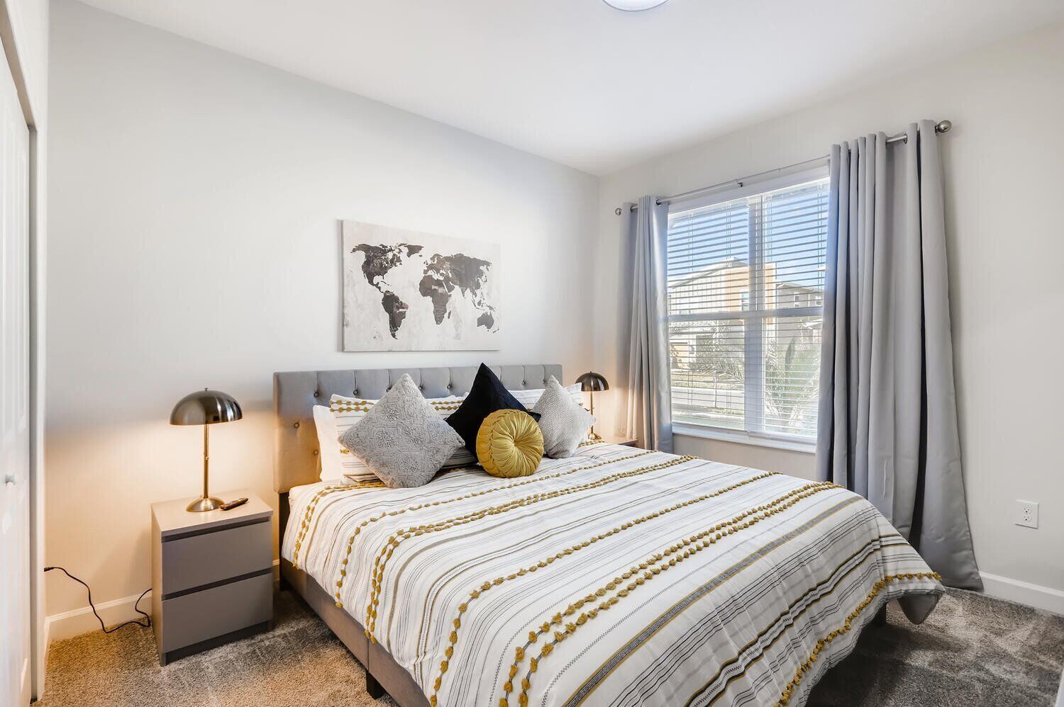 A neatly arranged bedroom with a bed, two bedside lamps, a world map artwork, and a window with curtains.