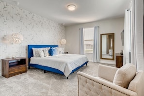 A bedroom with a bed, a nightstand, a chair, a mirror, and a window with curtains.