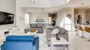 Ein großzügiger Wohnbereich mit Flachbildfernseher, grauen Sofas, einem blauen Sessel und einer Küche im Hintergrund.