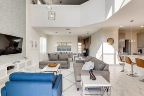 Ein geräumiger Wohnbereich mit Flachbildfernseher, grauen Sofas, einem blauen Sessel und einer Küche im Hintergrund.