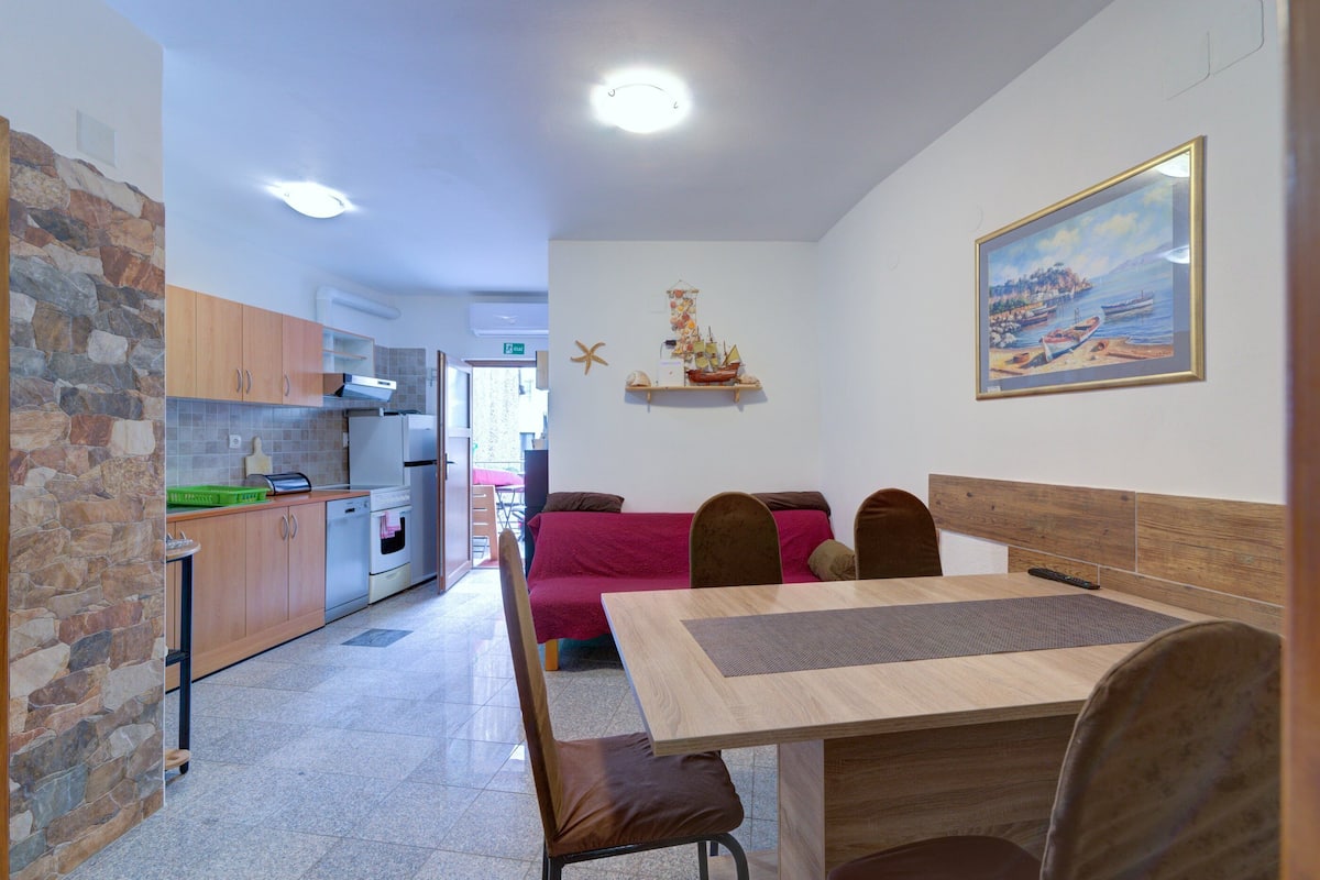 A compact kitchen with modern appliances, a dining area with a wooden table and chairs, and a cozy seating area with a red sofa.