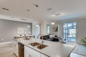 Una cocina moderna con una isla central, iluminación colgante y un comedor con sofá y mesa de centro.