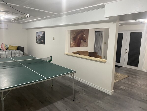 A room with a ping pong table, a couch with pillows, a mounted horse painting, and a door with a black shutter.