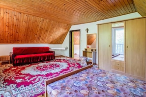 Una habitación con techo inclinado, un sofá rojo, un tapete estampado, una cama con colcha floral y un armario de madera.