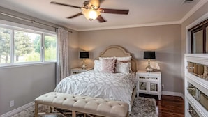 Een slaapkamer met een groot bed, twee nachtkastjes, een plafondventilator en uitzicht op het natuurlijke landschap door een raam.