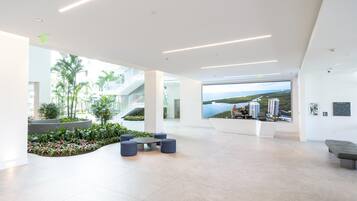 Un lobby moderno con un amplio ventanal que ofrece vistas a un paisaje urbano costero, un mostrador de recepción y una zona de estar con un pequeño jardín.