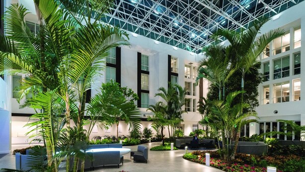 Un vaste atrium intérieur doté d’un haut plafond, avec un bâtiment moderne aux grandes fenêtres, entouré d’une végétation luxuriante et agrémenté d’espaces de détente.