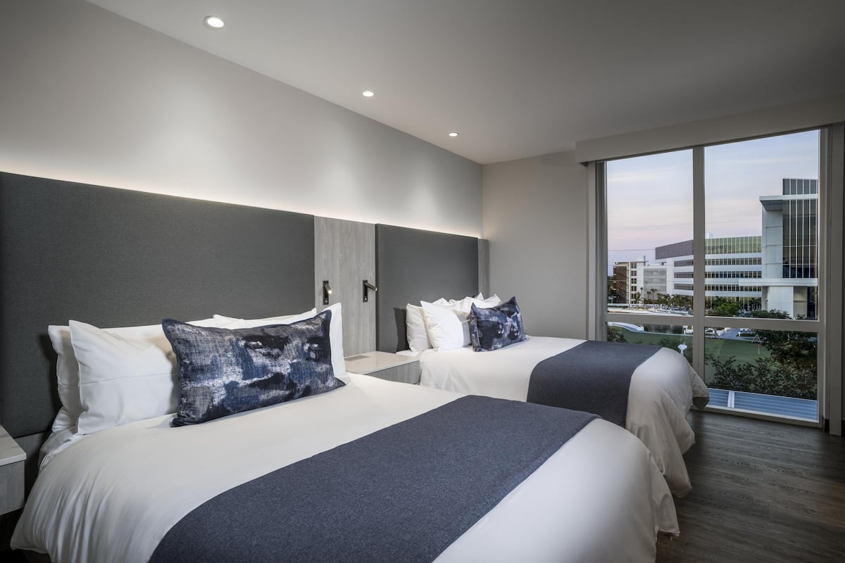 A modern hotel room with two beds, a large window with a city view, and a minimalist design.