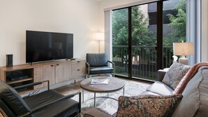 Un soggiorno moderno con una TV a schermo piatto, comodi posti a sedere, un tavolino da caffè e un'ampia porta scorrevole in vetro che dà su un balcone con vista sugli alberi.