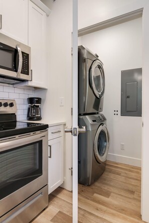 Compact kitchen with stainless steel oven, microwave, and coffee maker, featuring white cabinetry and a tiled backsplash.