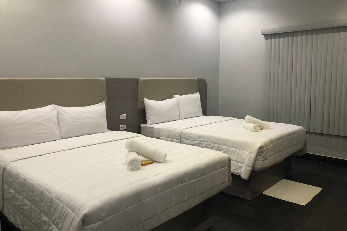 Habitación de hotel con dos camas, cada una con ropa de cama y almohadas blancas, un cabecero gris y una ventana con persianas.