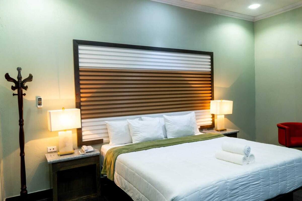 Habitación de hotel con cama, dos lámparas de noche, una silla roja y un teléfono fijo en la pared.