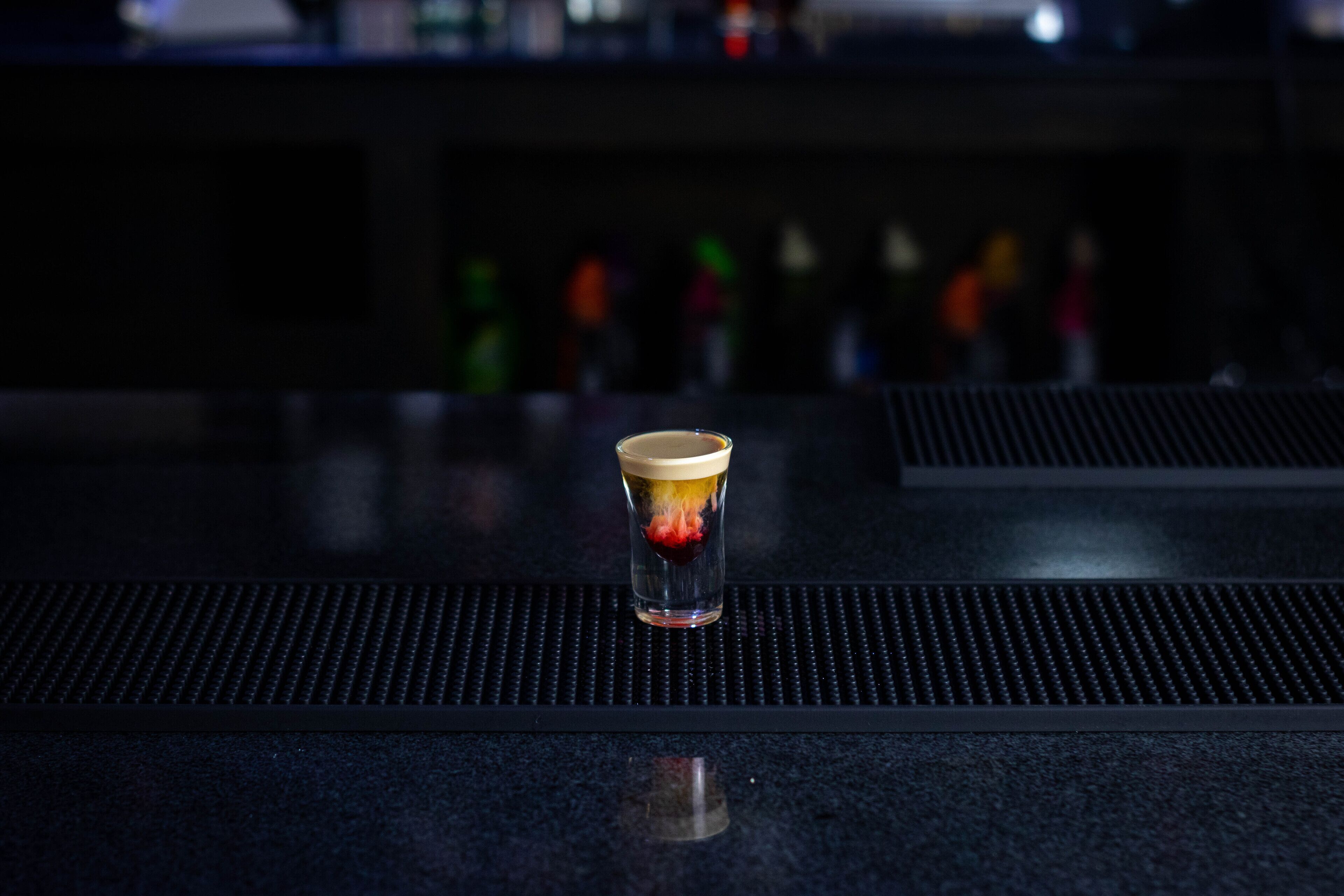A bar counter with a glass of beer and a bottle of beer in the background.