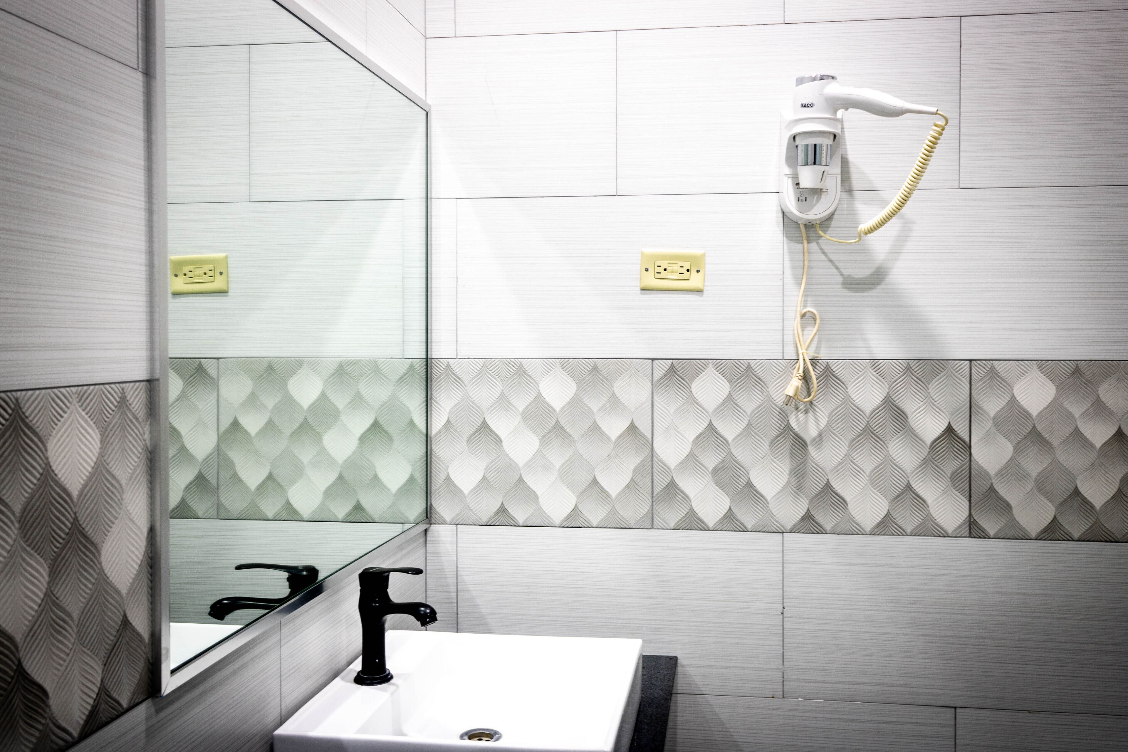 Ein Badezimmer mit einem weißen Waschbecken, einem schwarzen Wasserhahn, einem Spiegel und einem an der Wand befestigten Fön.