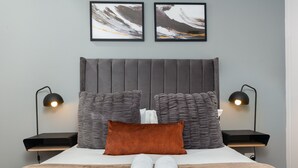 A neatly made bed with a grey headboard, two grey pillows, a brown pillow, and two white towels. There are two black bedside tables with lamps. Two framed abstract paintings hang on the wall above the bed. - Modern and Sophisticated 2 Bedroom Apartment (Cape Town)