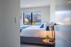 A modern bedroom with a large bed, a view of mountains, and a small bedside table with a lamp and a plant.