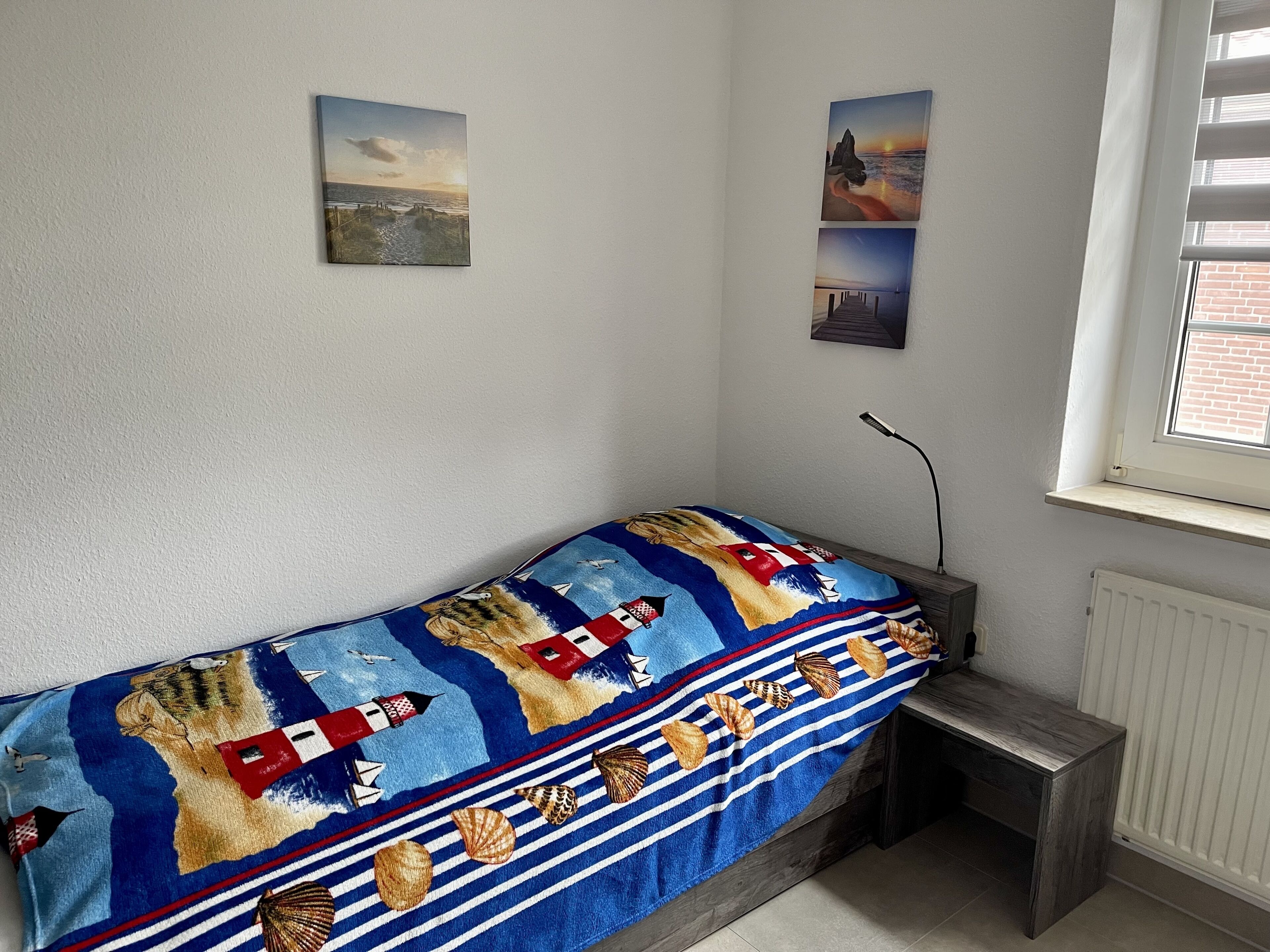 A small, single-bed room with a nautical-themed bedspread, a window with blinds, and two framed pictures on the wall.